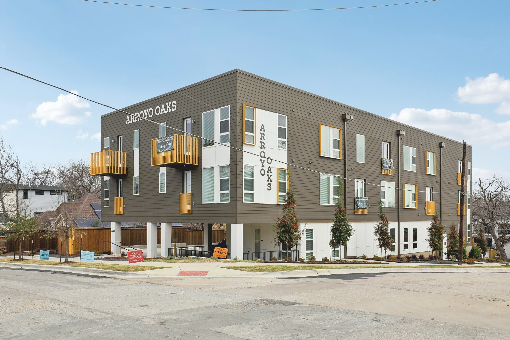 Modern three-story multifamily apartment building in Dallas, Texas with elevated podium structure, yellow balcony accents, and contemporary exterior siding at Arroyo Oaks.